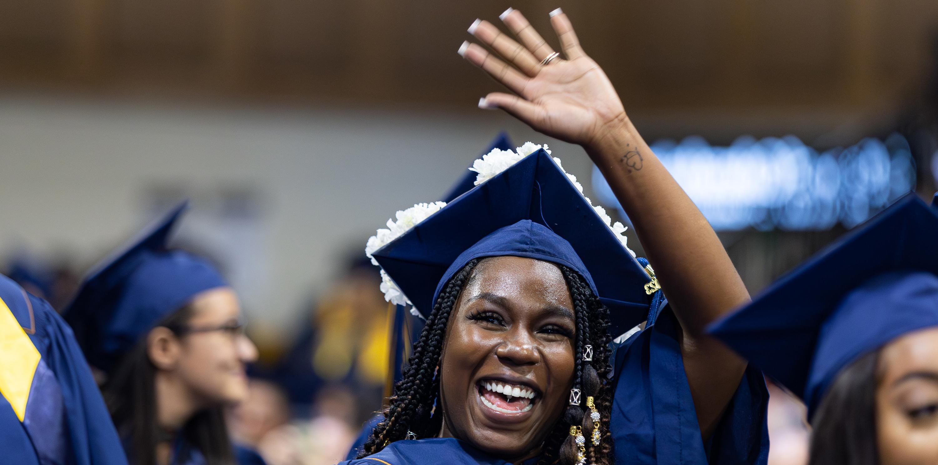 Female college student in her cap and gown at graduation, smiling and waving